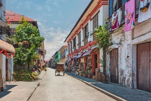 A cobblestone street lined with historic Spanish-style buildings adorned with balconies, colourful awnings, and vibrant textiles hanging, with a horse-drawn carriage in the distance under a sunny sky.