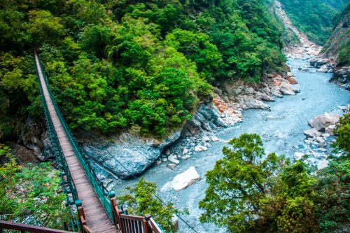 A high angle shot of a modern suspension footbridge spanning a steep, densely forested gorge with blue flowing water underneath