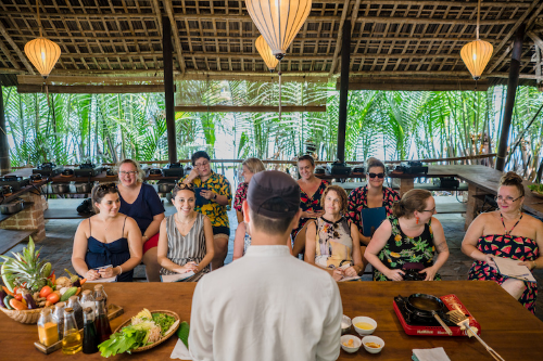  People seated at a long table in a tropical setting, attending a cooking class led by an instructor.