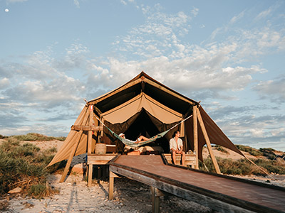 A woman lays in a hammock while a man sits on the porch of their glamping tent by the beach.