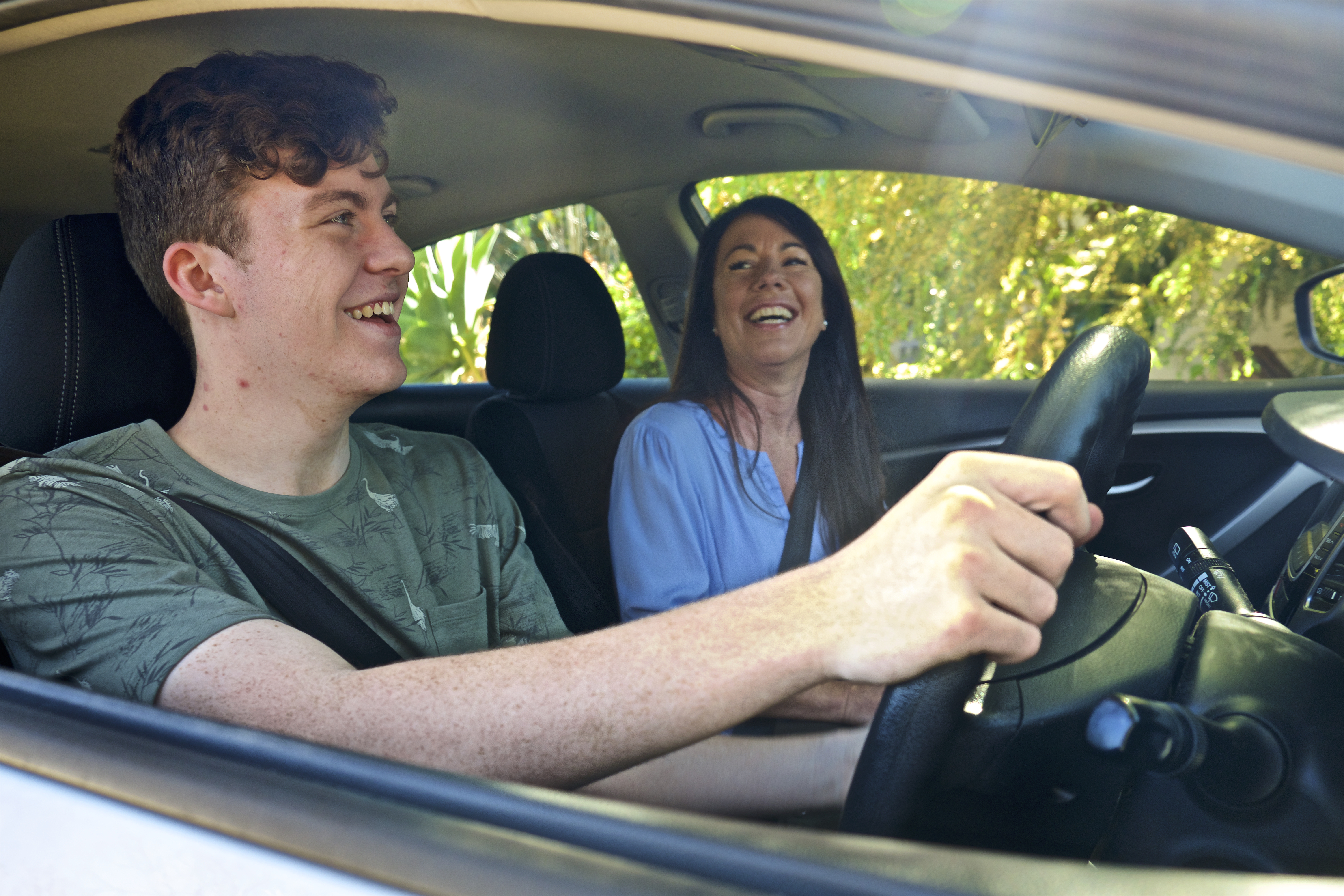 Young person learning to drive with an adult passenger, both smiling and enjoying the driving lesson inside a car during the day