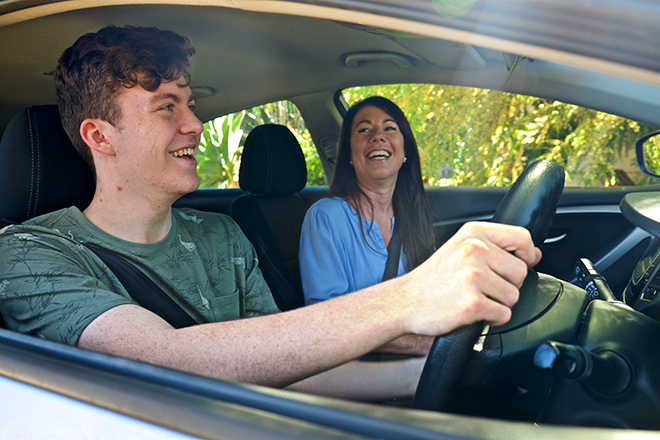 Young person learning to drive with an adult passenger, both smiling and enjoying the driving lesson inside a car during the day